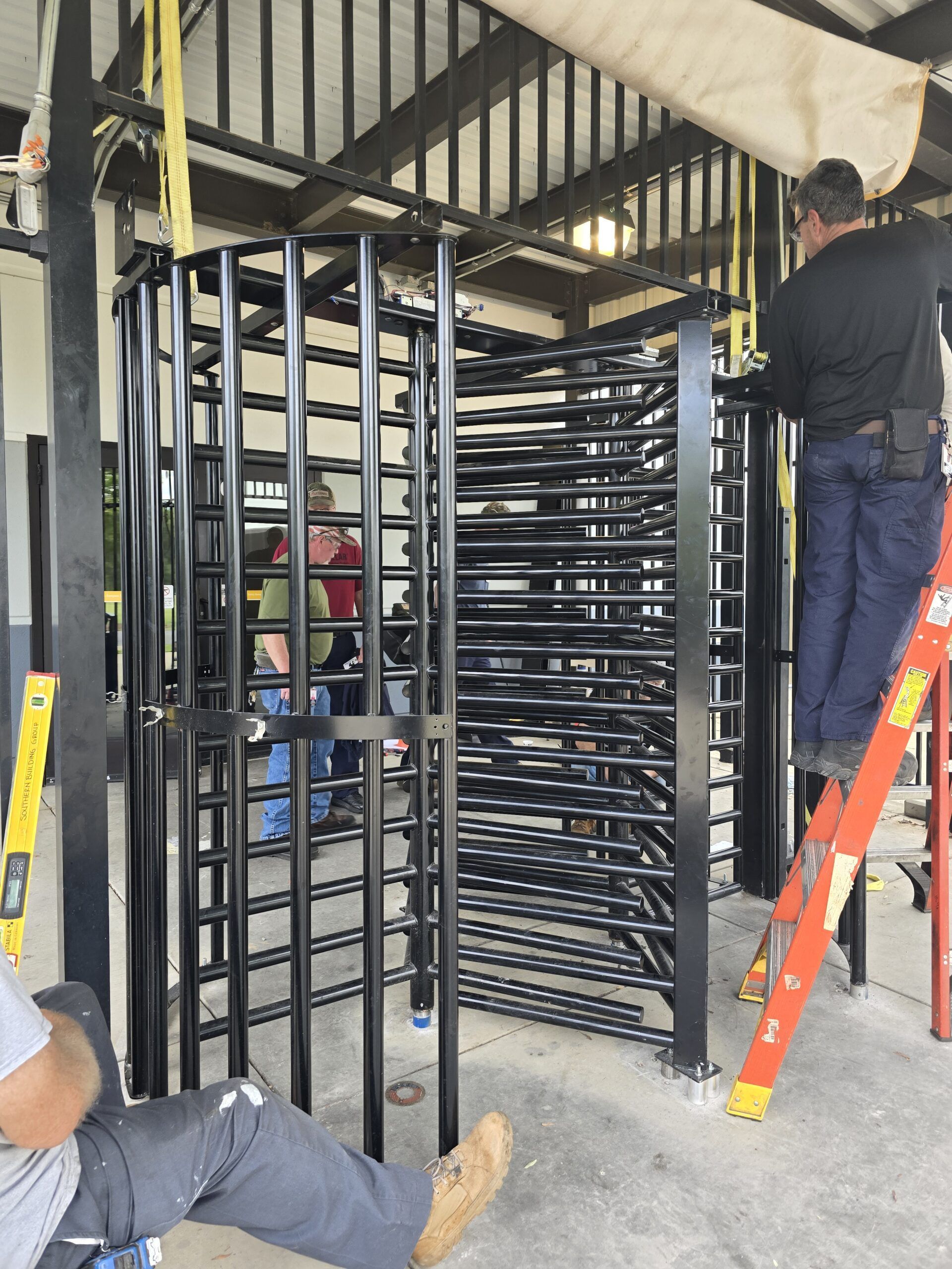 Workers installing a black metal turnstile in a building. One worker on a ladder.