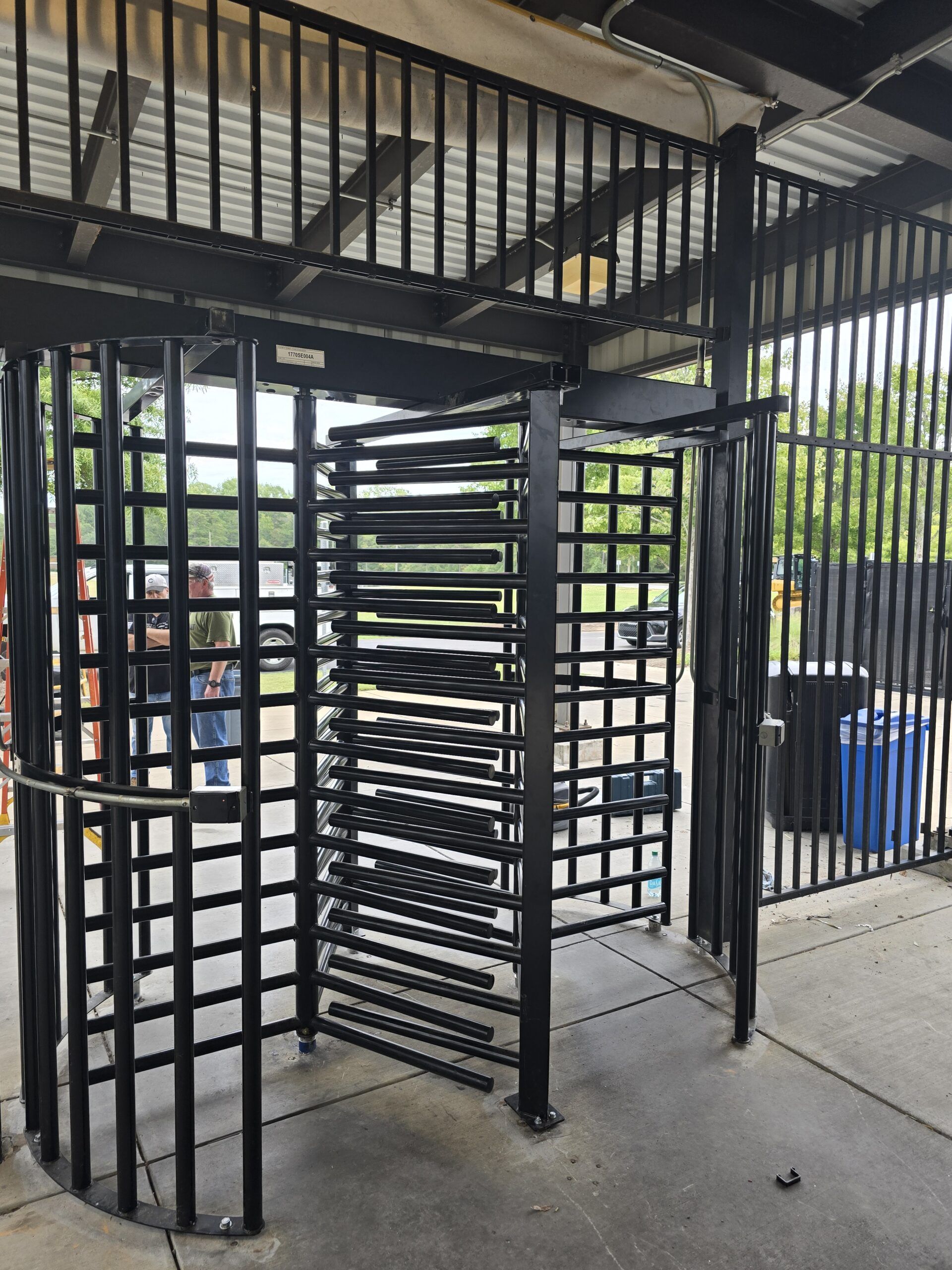 Black metal turnstile at an entrance to a gated area. Open passageway on one side.