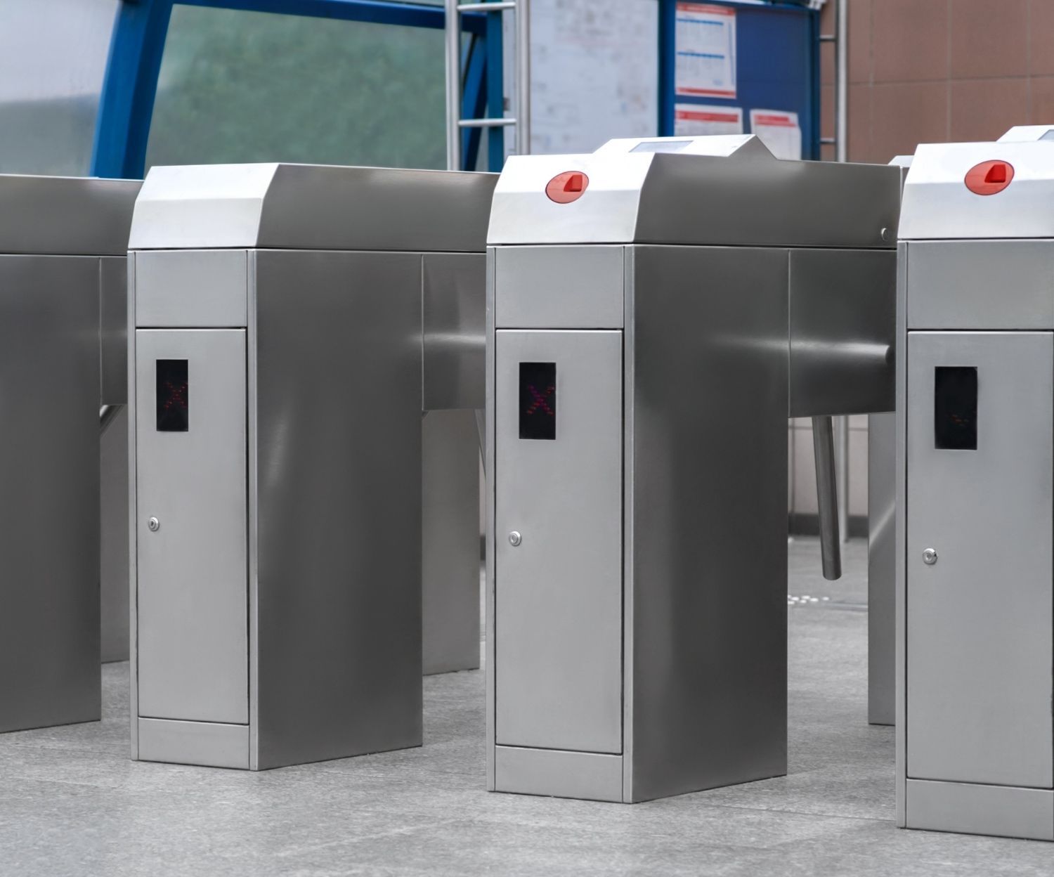 Turnstiles, metal, with red indicator lights, in a public space Turnstiles in a transit station. Stainless steel rectangular structures. Red button on top. Dark display panel on front.