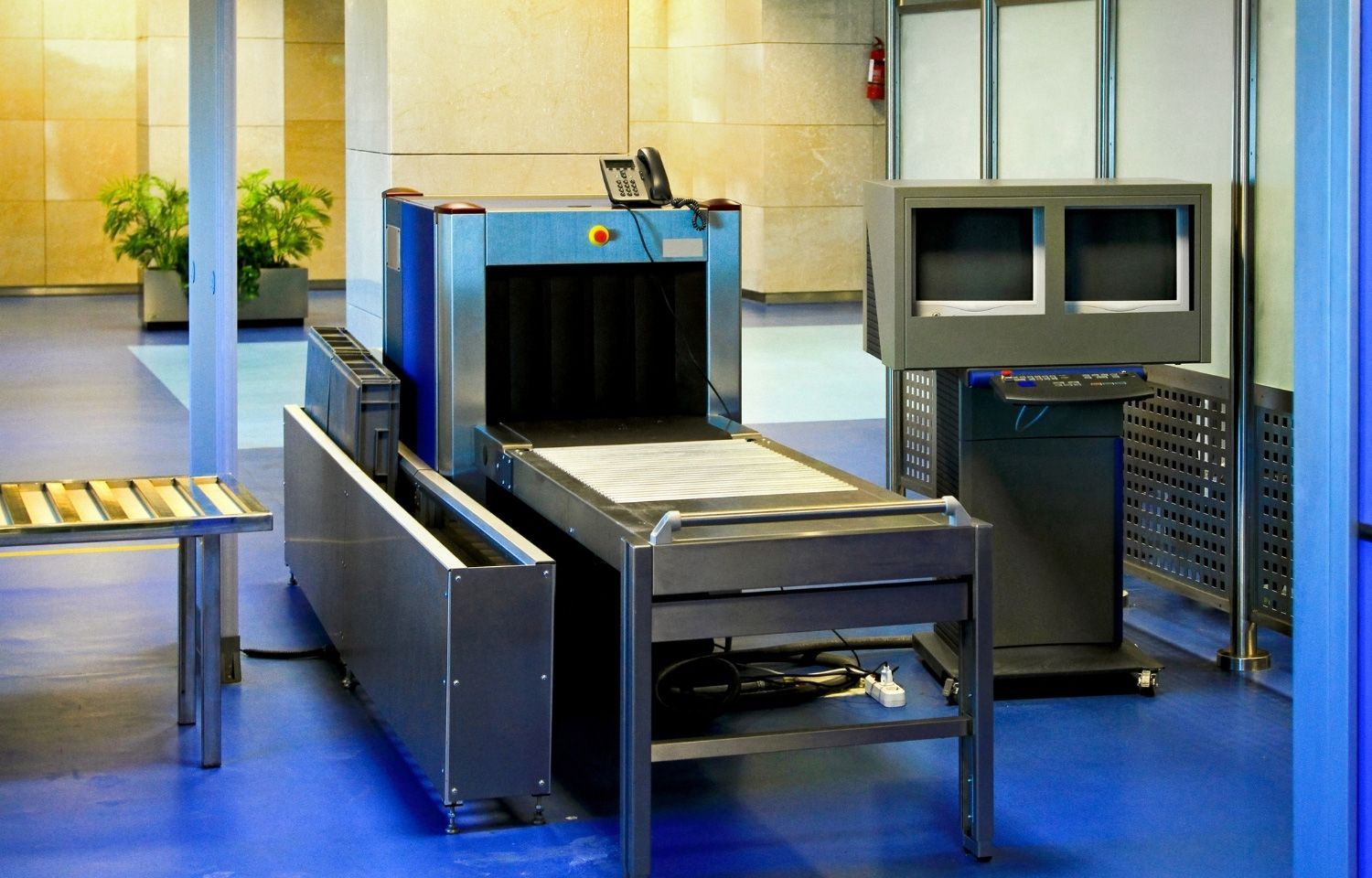 Airport security X-ray scanner with conveyor belt, stainless steel, and monitor. Blue walls, tiled floor.