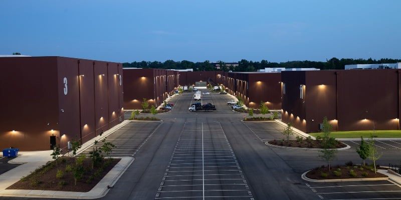 Rows of brown buildings with lights, a long gray road, and blue sky An aerial view of a brown industrial building complex at dusk with parking and landscaping.