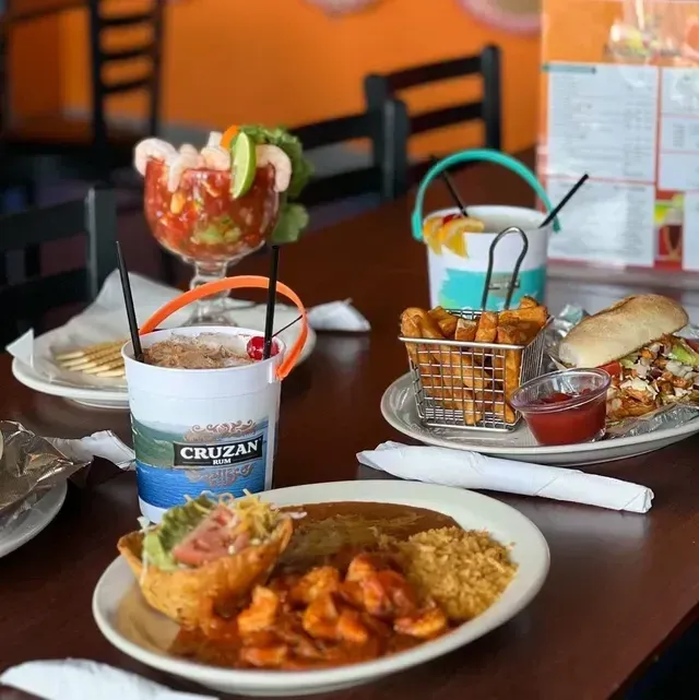 Table of Mexican food: shrimp cocktail, cocktails in buckets, fries, sandwich, and plate of food.