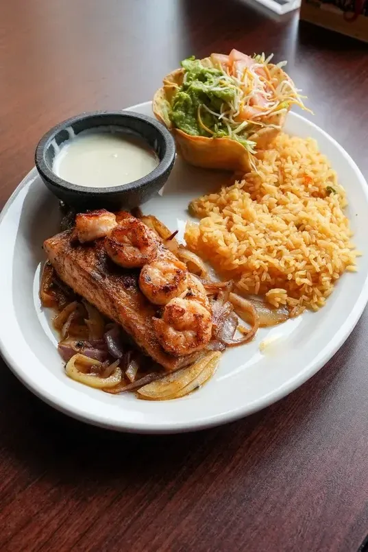 Grilled salmon and shrimp over onions, with rice, guacamole, and a sauce on a plate.