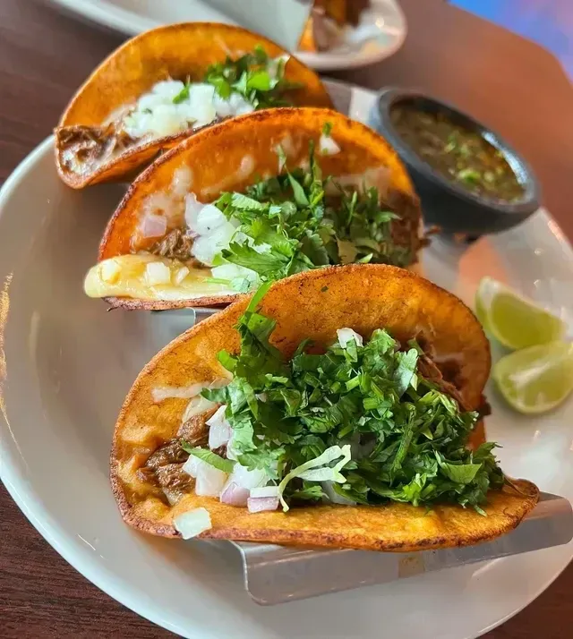 Three tacos in a metal stand on a white plate, topped with cilantro and onions, with salsa and lime wedges.