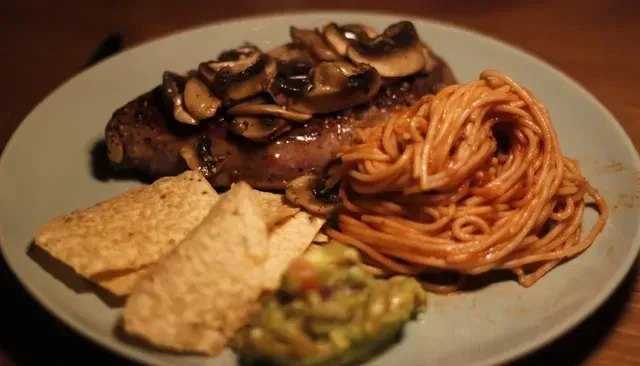 Steak with mushroom topping, spaghetti, tortillas, and guacamole on a plate.