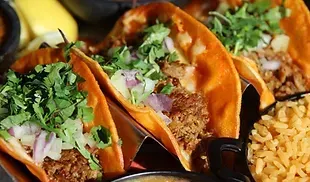 Close-up of three tacos in orange shells, filled with meat and topped with cilantro and onions, with a side of rice.