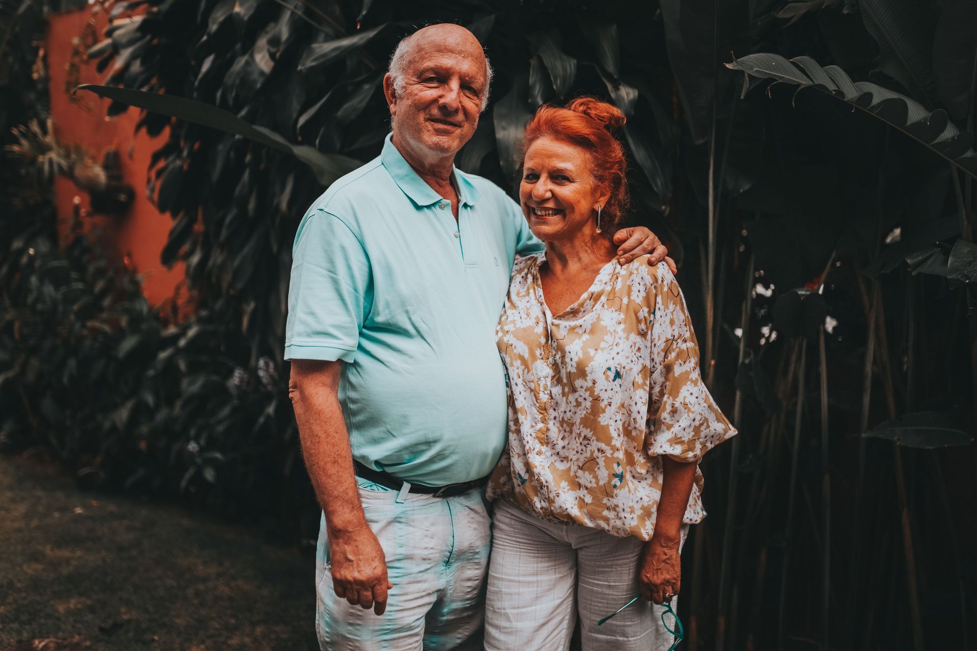 Smiling couple standing together in front of lush green foliage, wearing light-colored clothes
