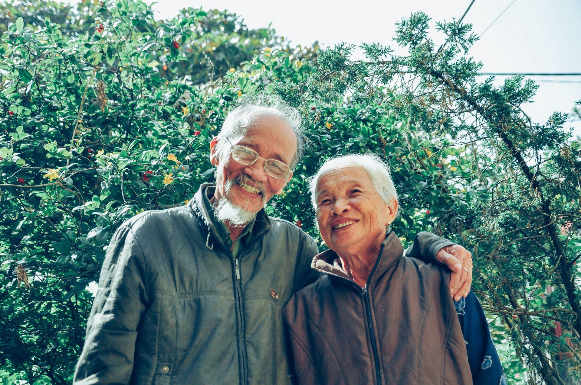 Two smiling people standing together outdoors in front of lush green trees.