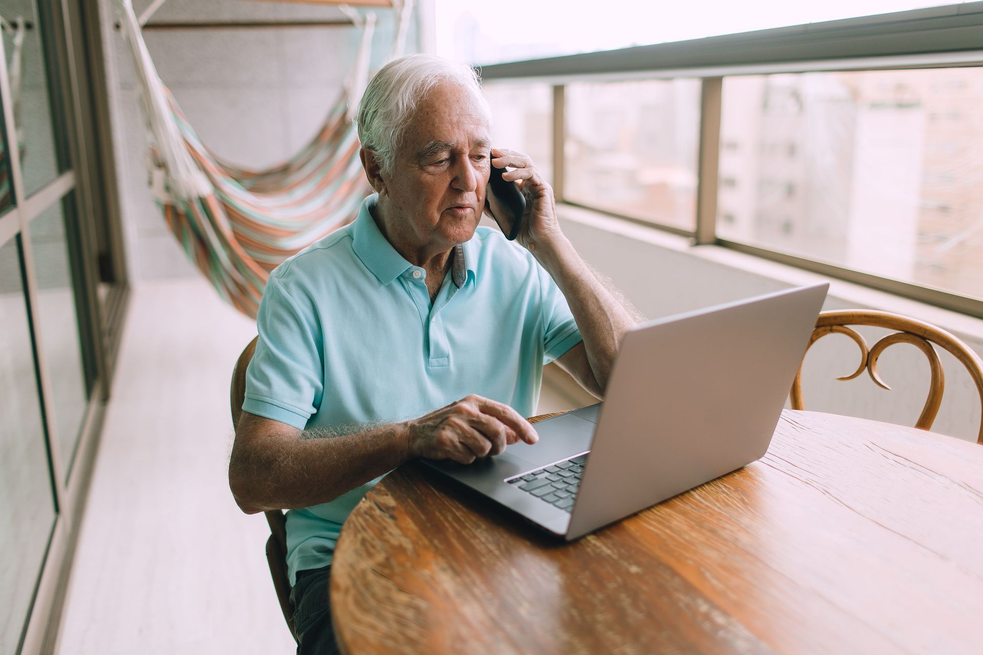 Man using a laptop and phone at a wooden table near a bright window and hammock chair