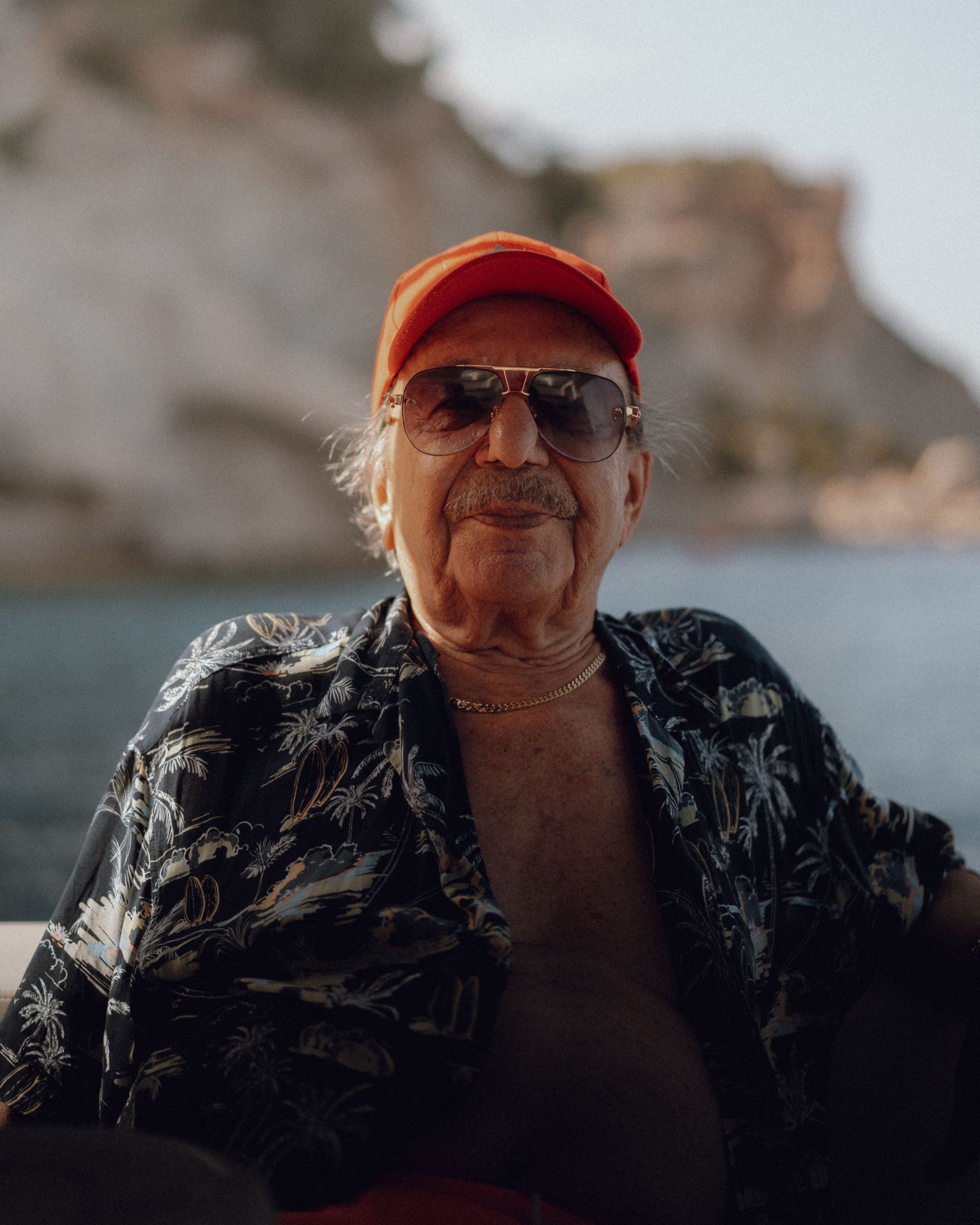 Person wearing a red cap and sunglasses sits by the sea with rocky cliffs in the background.