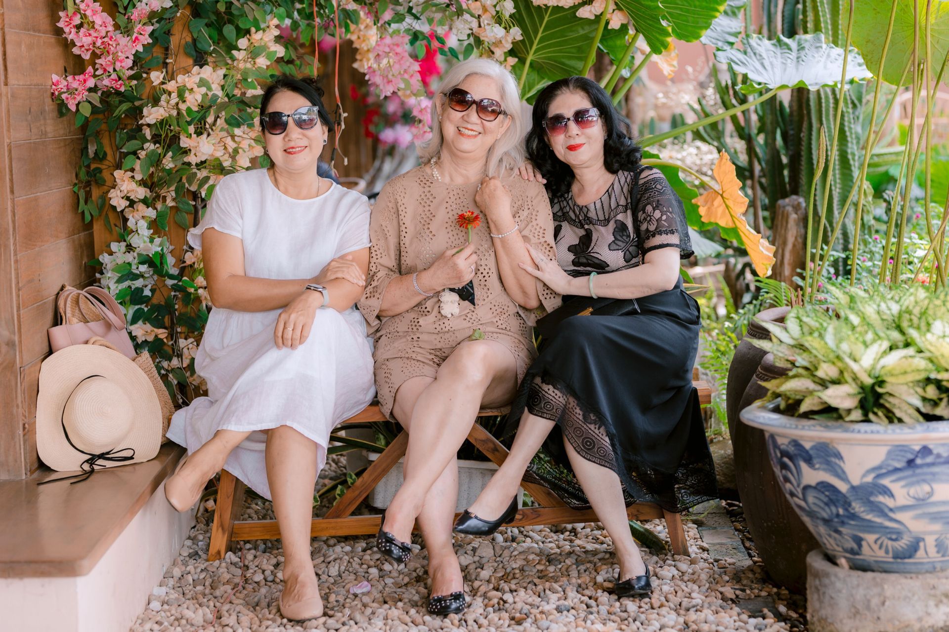 Three women seated on a bench in a lush garden, smiling and posing among flowers and plants.