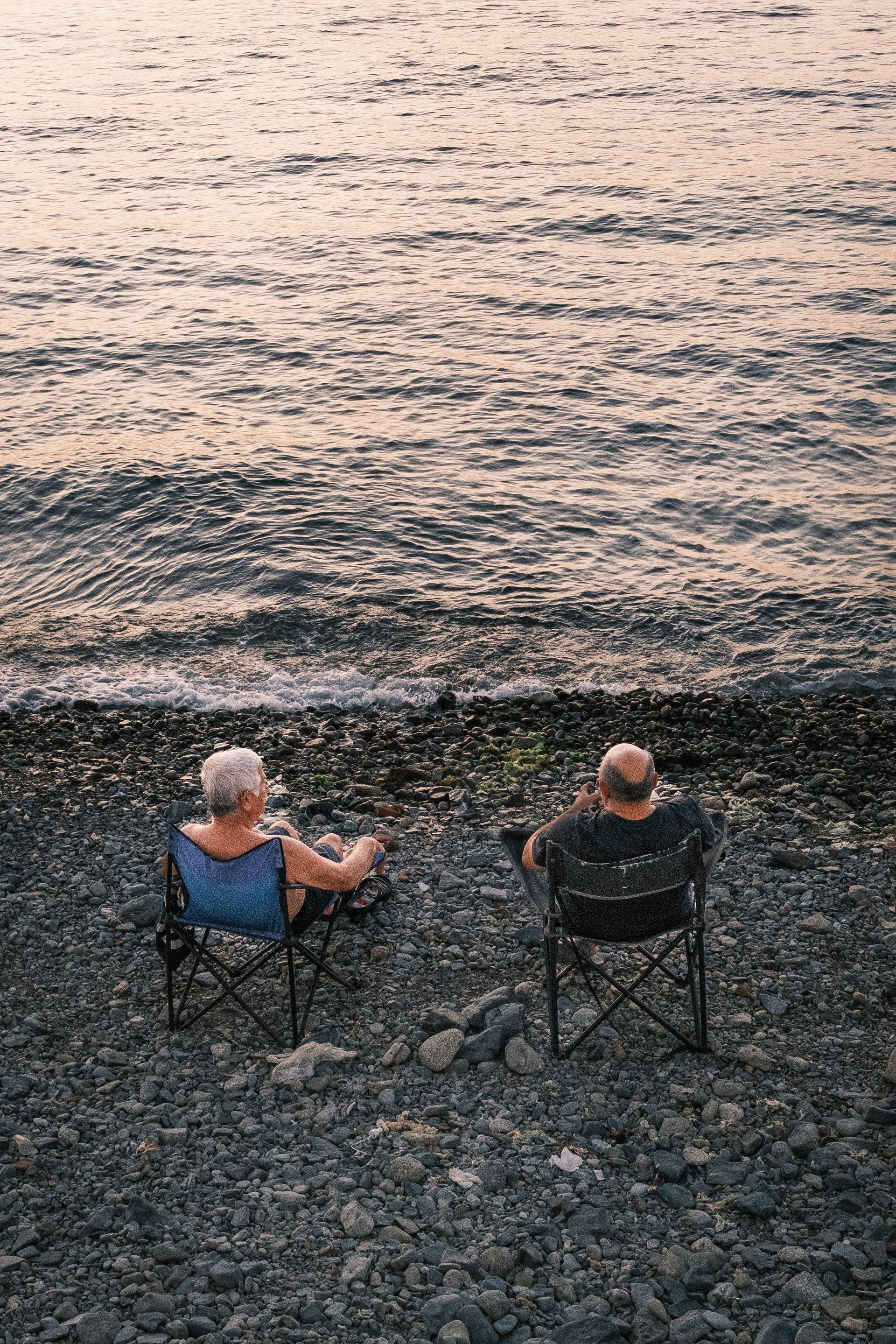 Two people in folding chairs facing a calm shoreline at dusk