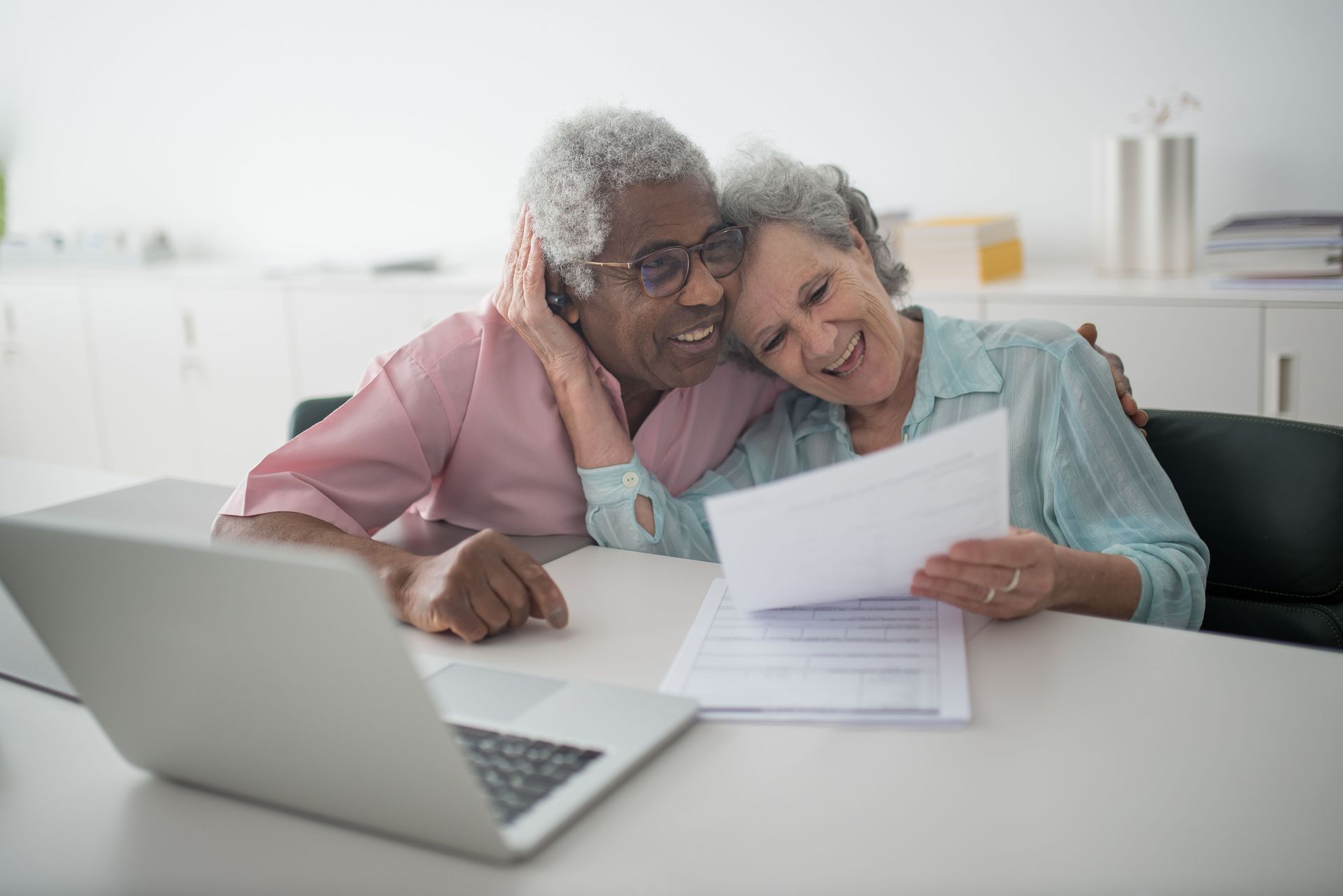 Two people at a desk reviewing papers beside an open laptop, smiling in a bright office