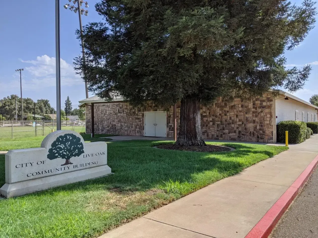 Stone monument sign beside a large tree and sidewalk at a park or civic building entrance