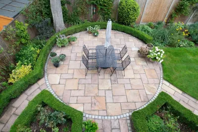 Backyard patio with a circular stone seating area, metal chairs, and a central fire pit surrounded by green garden beds