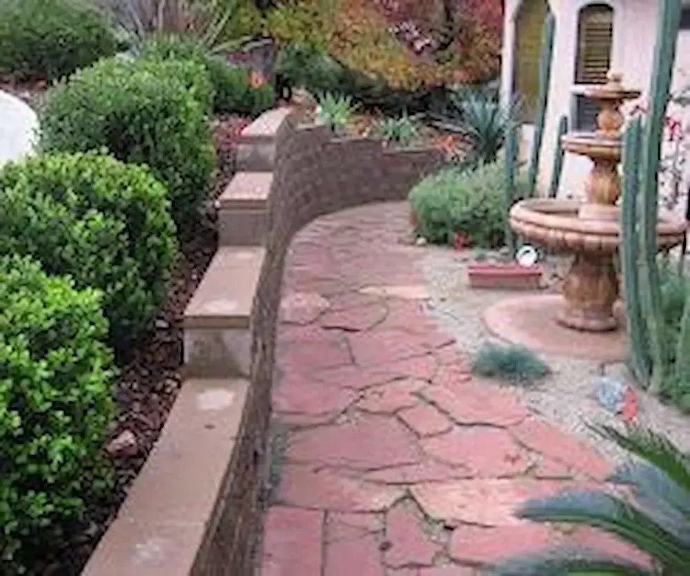 Curved garden walkway with pink stone pavers, trimmed hedges, and a stone fountain beside a house