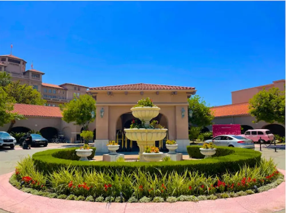 Decorative fountain in a landscaped courtyard with flowers, parked cars, and clear blue sky