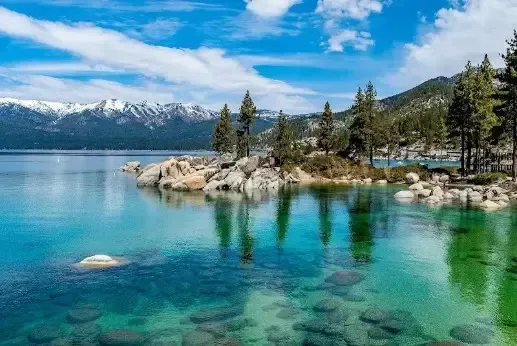 Turquoise mountain lake with rocky shore, pine trees, and snow-capped peaks under a clear sky