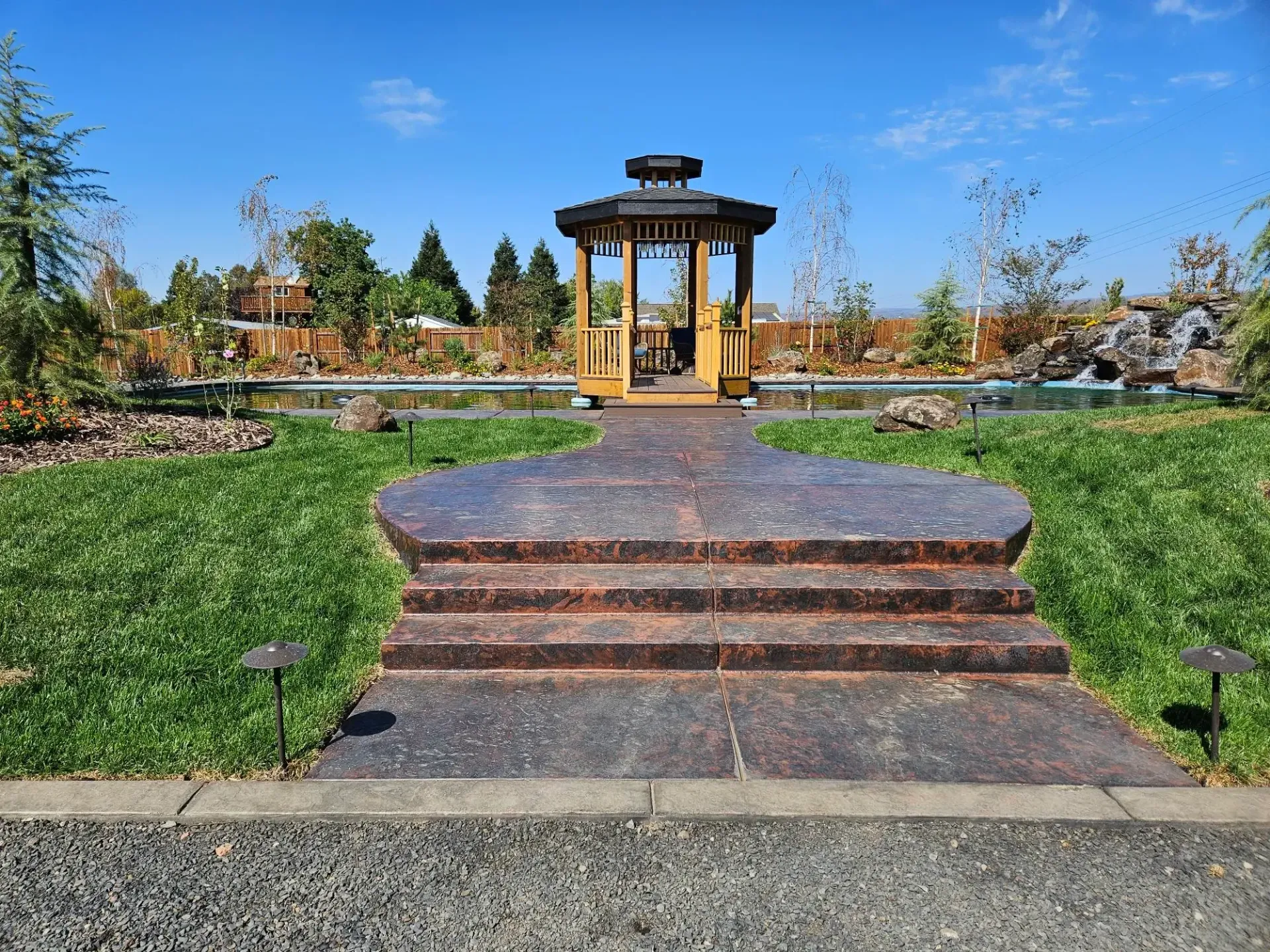 Stone steps lead to a small wooden gazebo in a landscaped garden under a blue sky