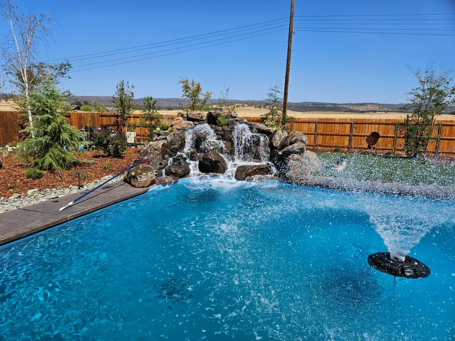 Backyard pool with blue water, rock waterfall, and desert landscaping under a clear sky