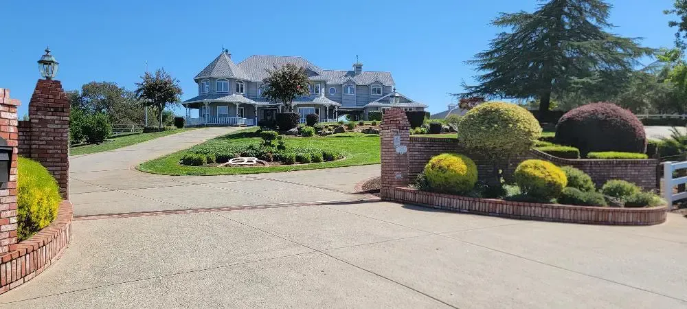 Mansion at top of a driveway, flanked by manicured shrubs and brick pillars under a clear blue sky