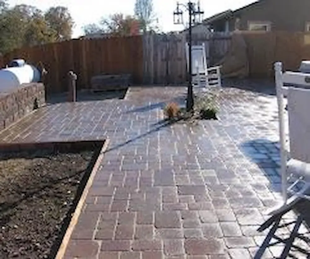 Brick-paved backyard patio with a raised planter and wooden fence in bright sunlight