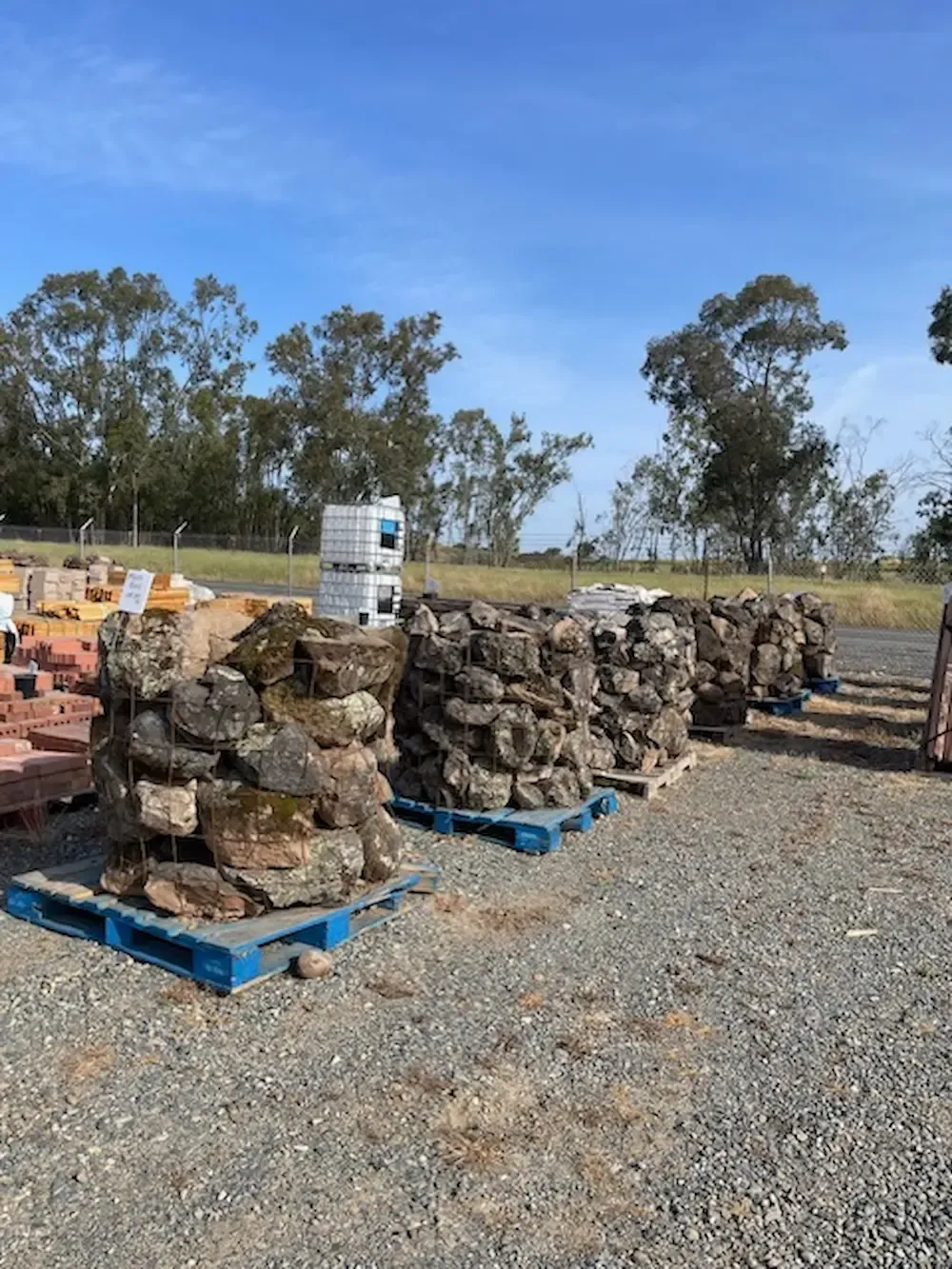 Blue pallets stacked with rough stone blocks outdoors on gravel, with trees and clear sky in the background
