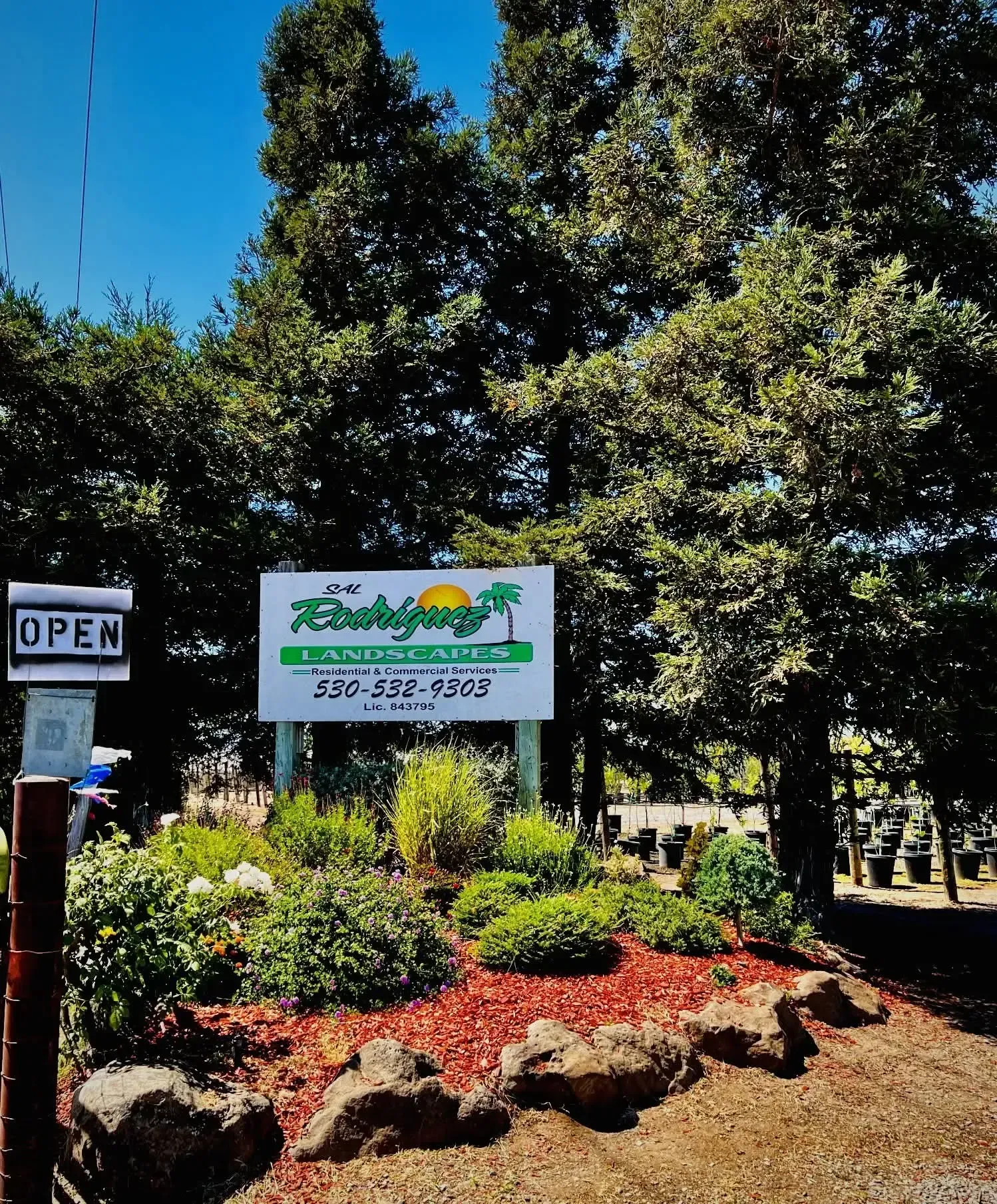 Outdoor garden entrance sign with trees, flowers, and an “OPEN” sign on the left.