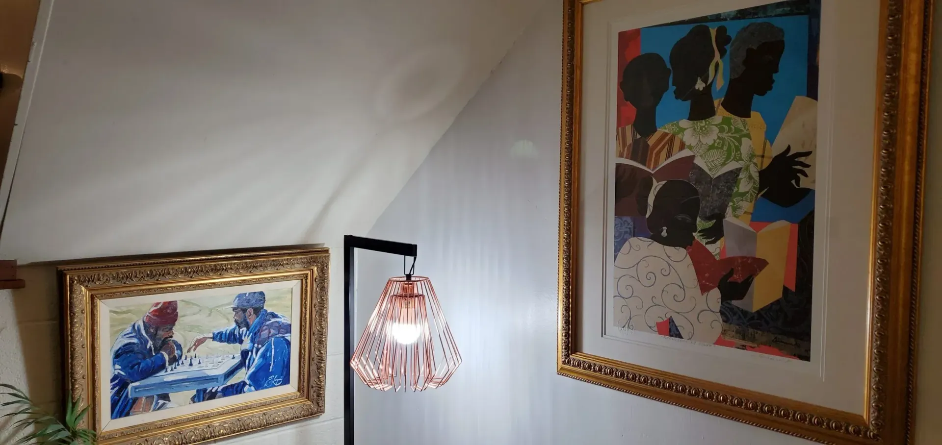 Two framed paintings hang on a white wall, a lamp with a pink shade is in between.