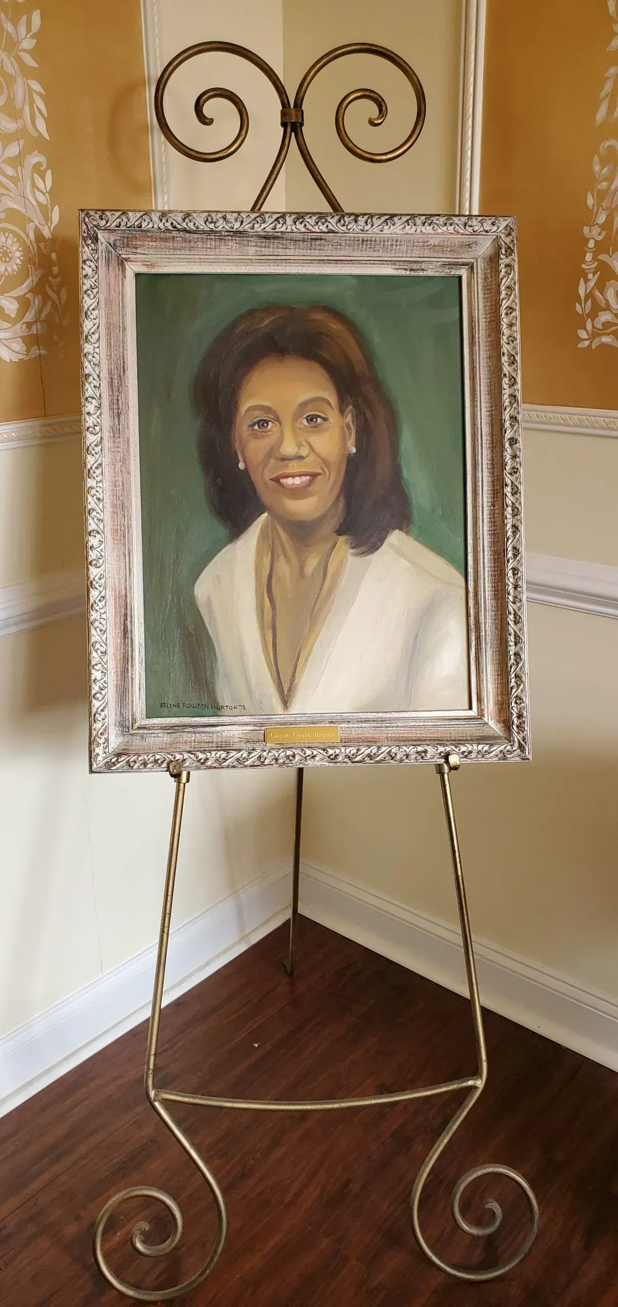 Portrait in a decorative frame on a golden easel in a corner. The painting is of a woman in a white top with green background.