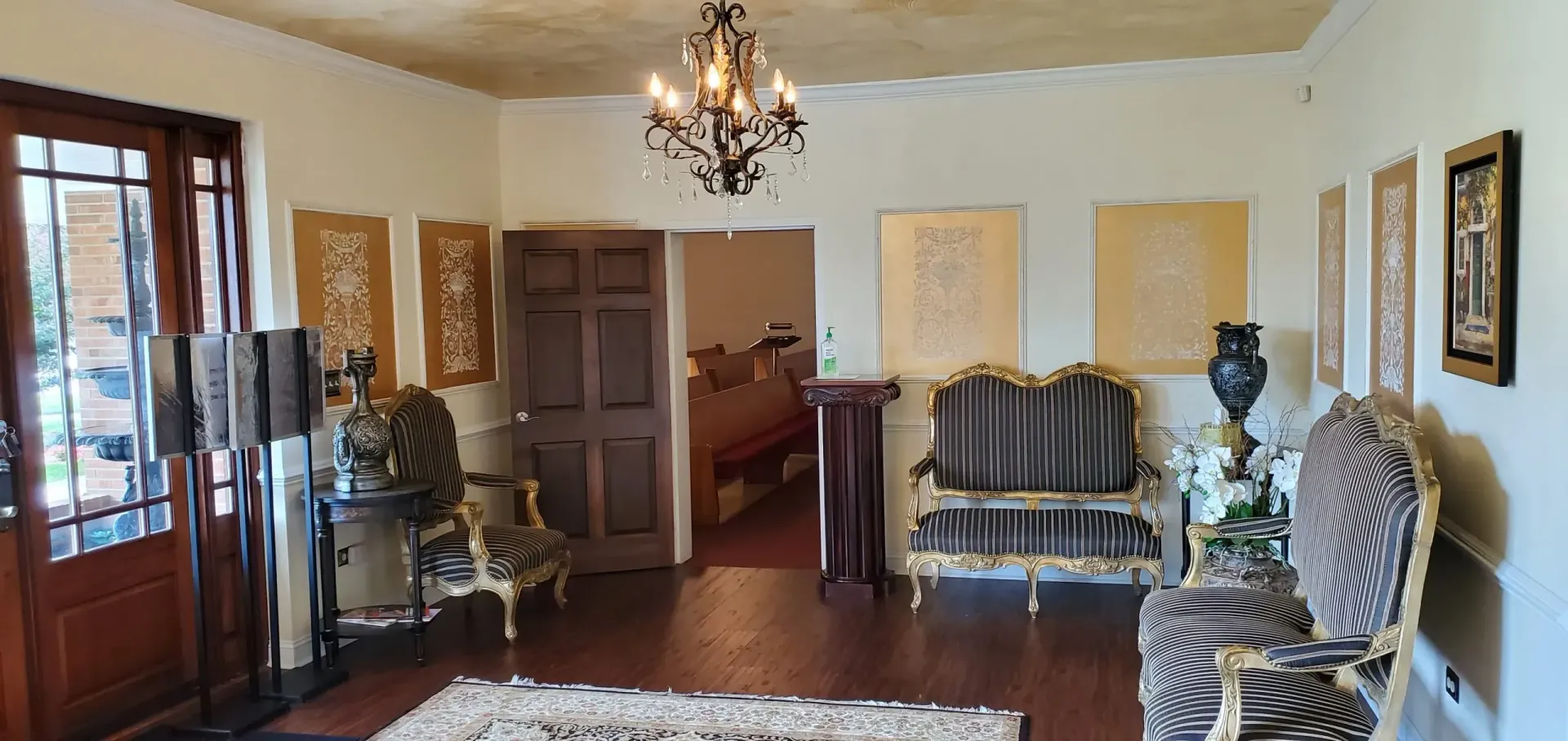 An ornate living room with chandeliers, sofas, and artwork. A wooden door and large windows are on the left.