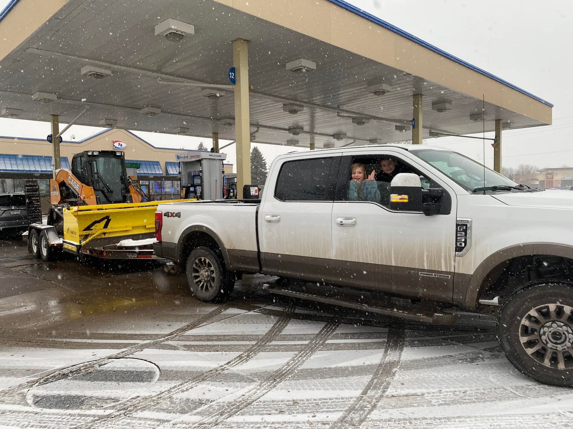 A truck is towing a plow in the snow at a gas station.