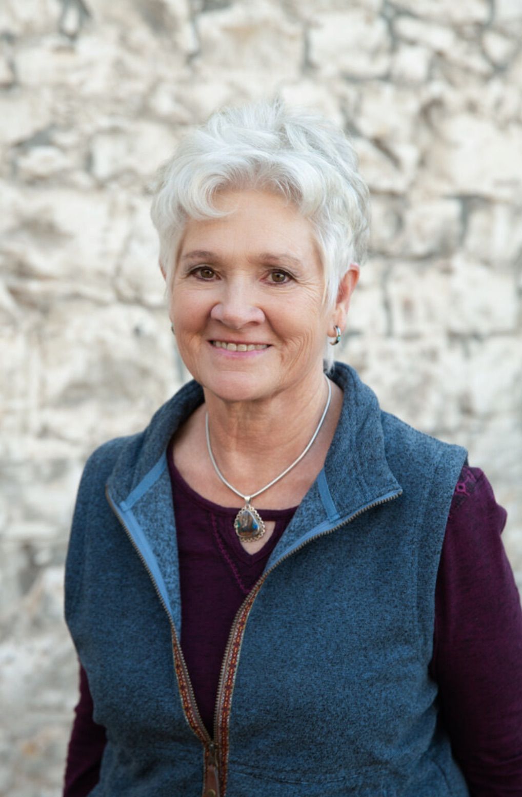 A woman wearing a blue vest and a necklace is standing in front of a stone wall.