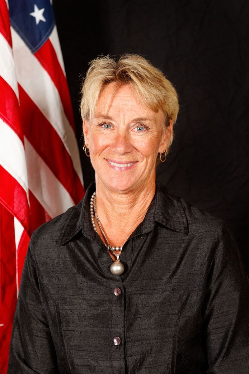 A woman in a suit is smiling in front of an american flag.
