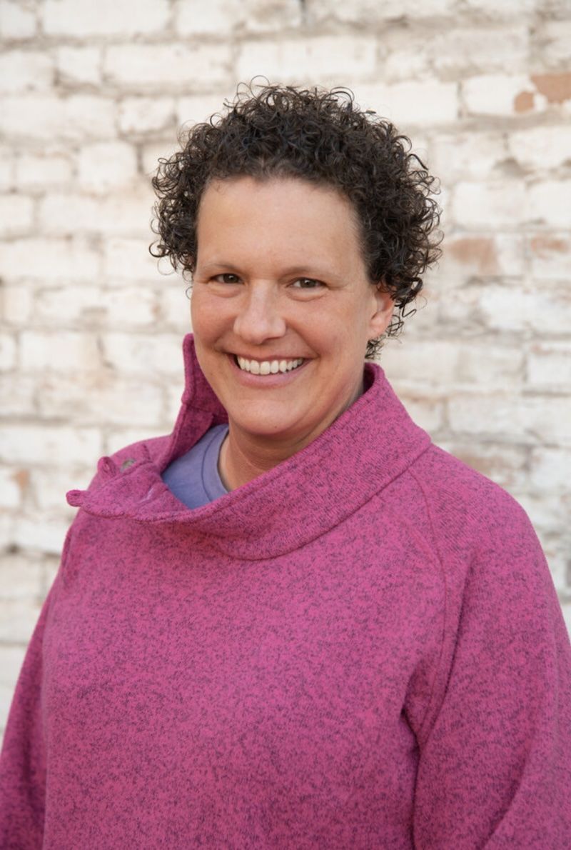 A woman in a purple sweater is smiling in front of a white brick wall.