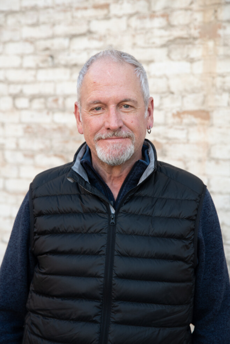 A man in a black vest is standing in front of a white brick wall.