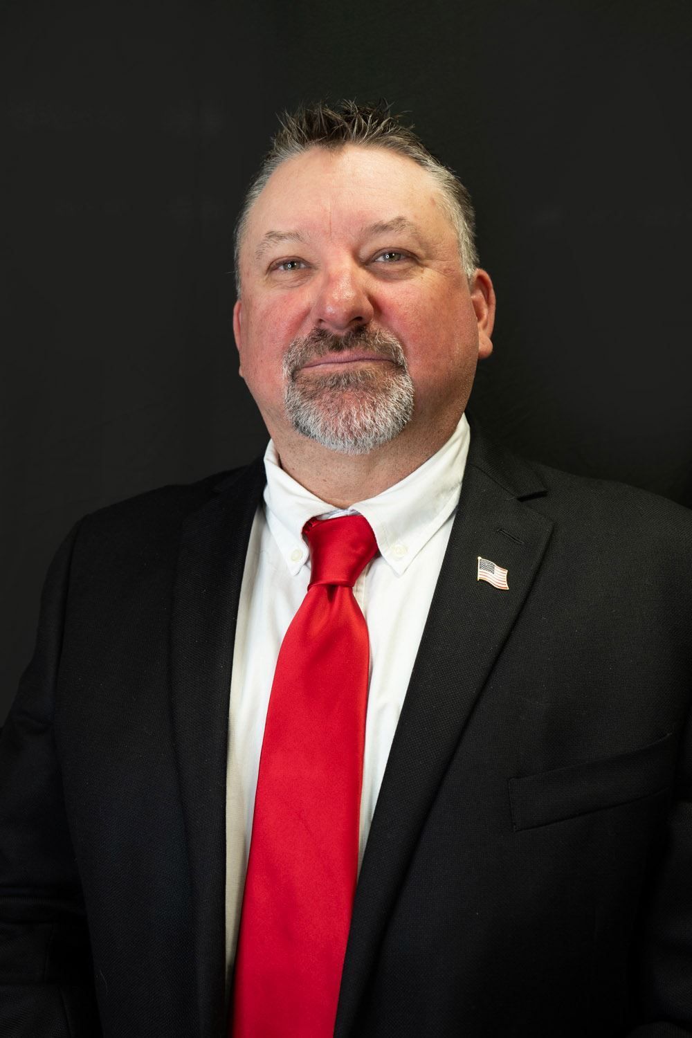 A man with a beard is wearing a uniform and tie in front of an american flag.