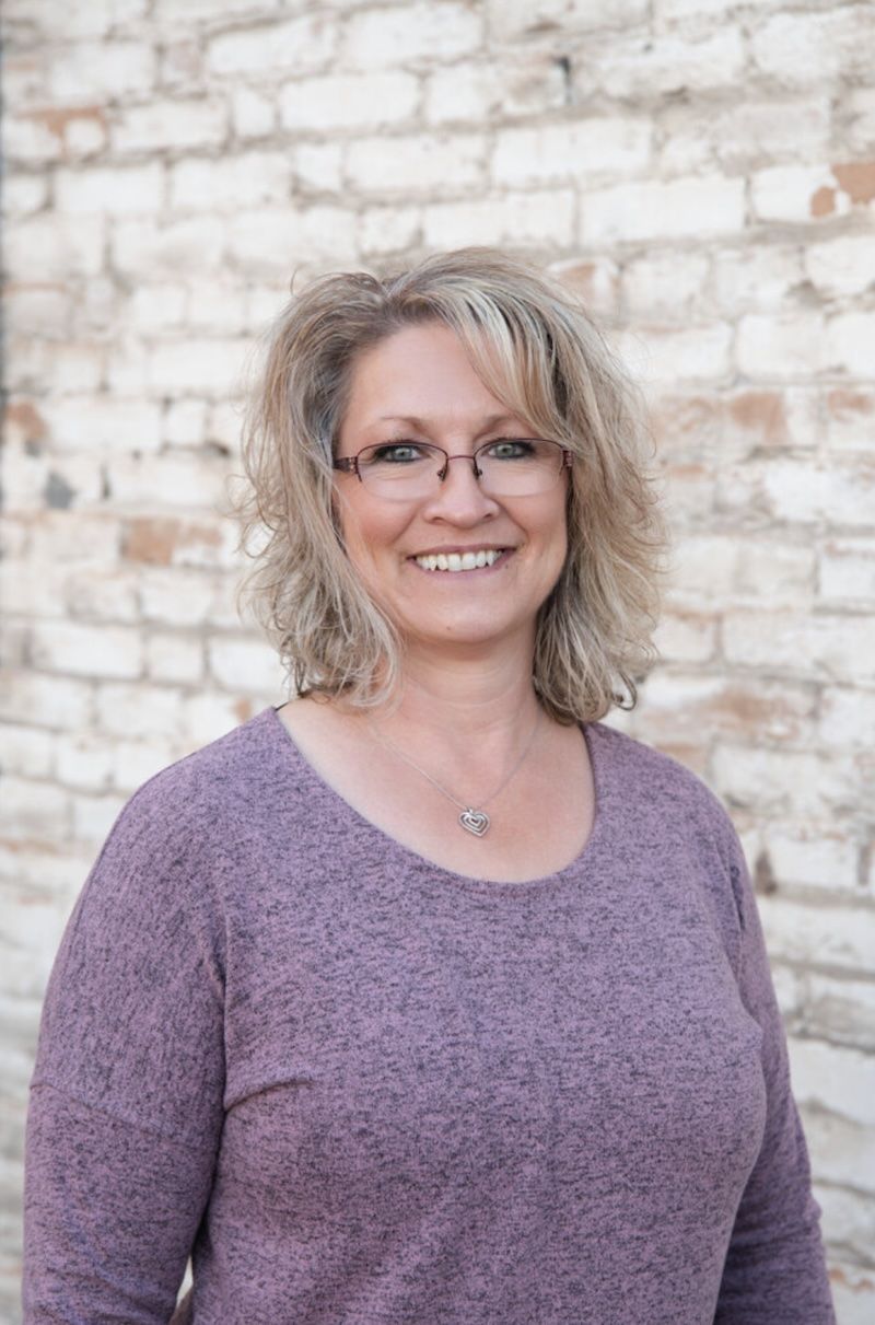 A woman wearing glasses and a purple shirt is smiling in front of a brick wall.