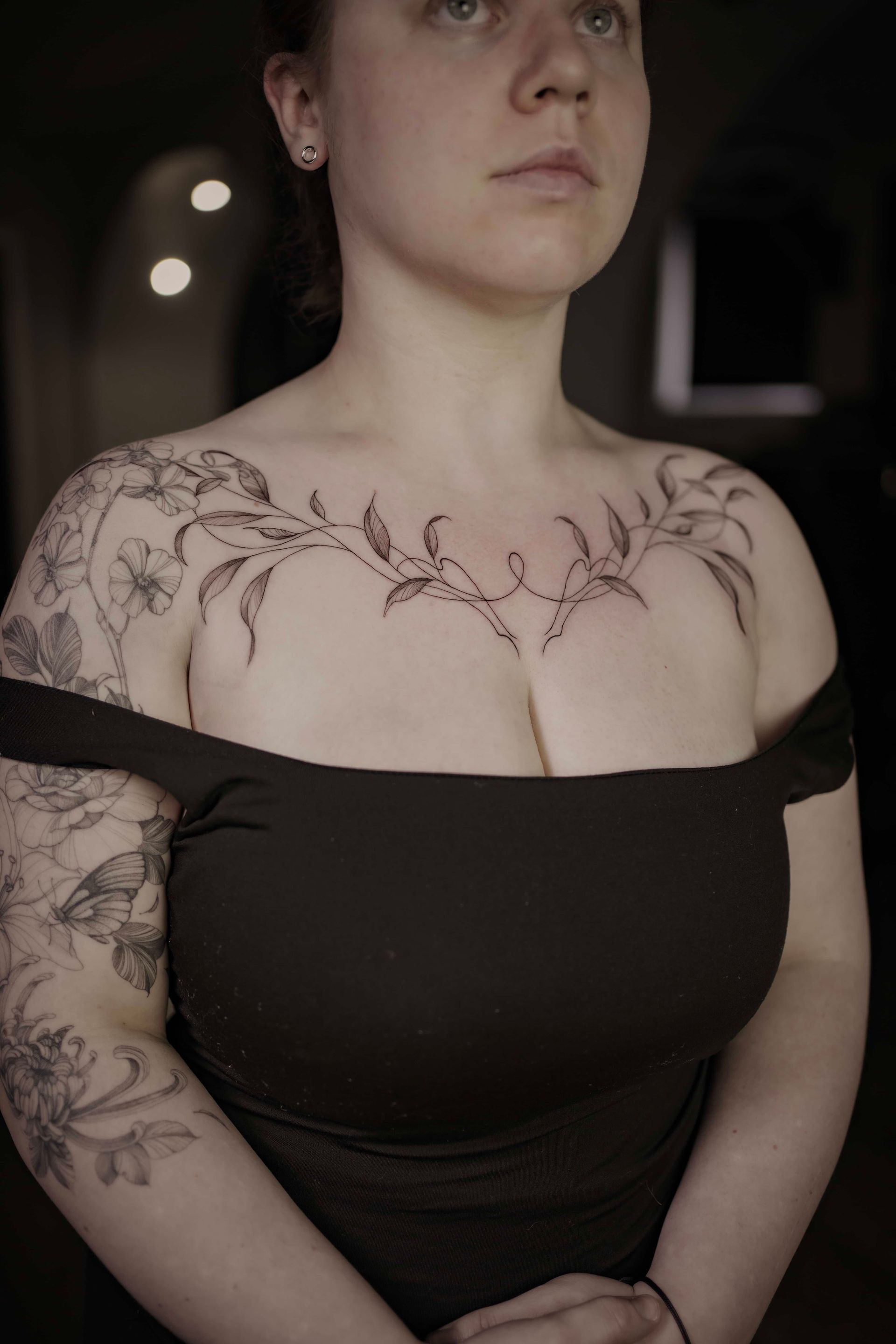 Woman with chest and arm tattoos, wearing a black off-shoulder top, indoors.