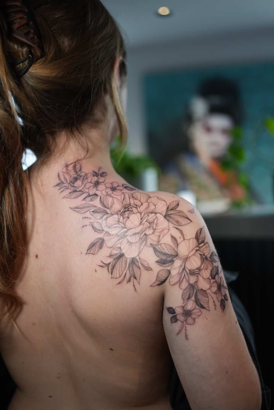 Woman's back and shoulder with a floral tattoo. The tattoo is black and grey. A mirror reflects a person in the background.