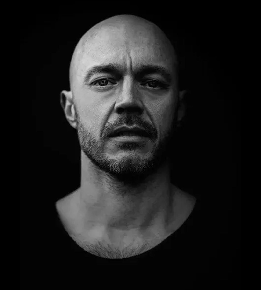 Bald man with a trimmed beard and somber expression, against a dark background.