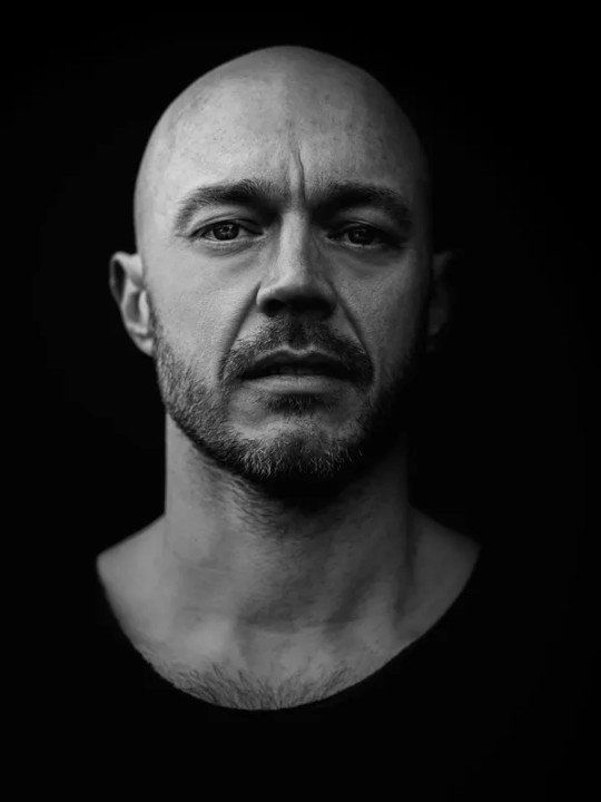 A black and white photo of a bald man with a beard looking at the camera.