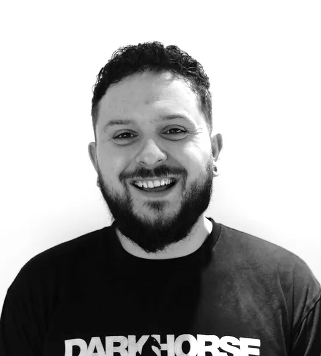 Smiling person with dark hair and beard, wearing a black t-shirt with 
