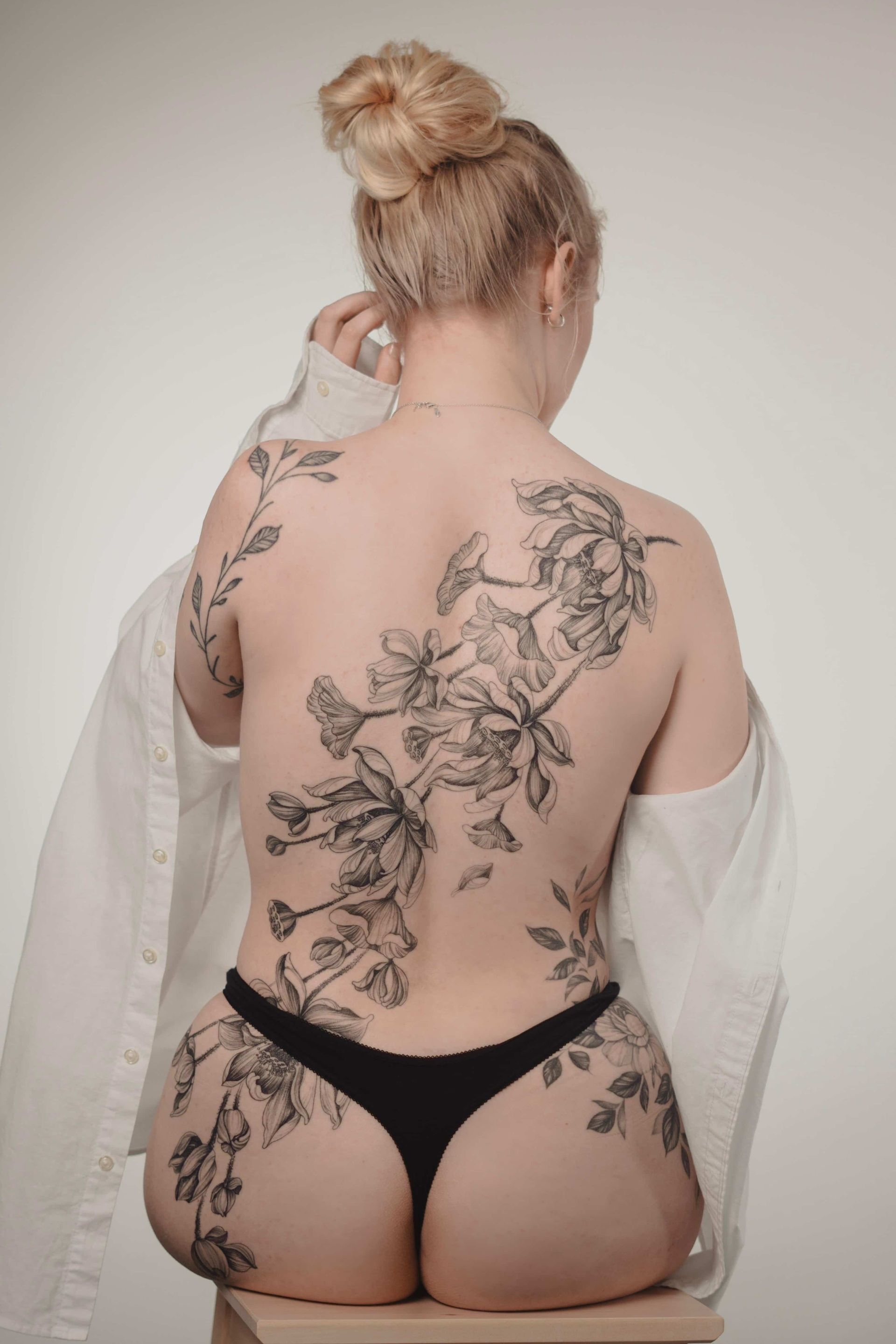 Woman with floral back tattoos, wearing a white shirt and black underwear, sitting on a stool.