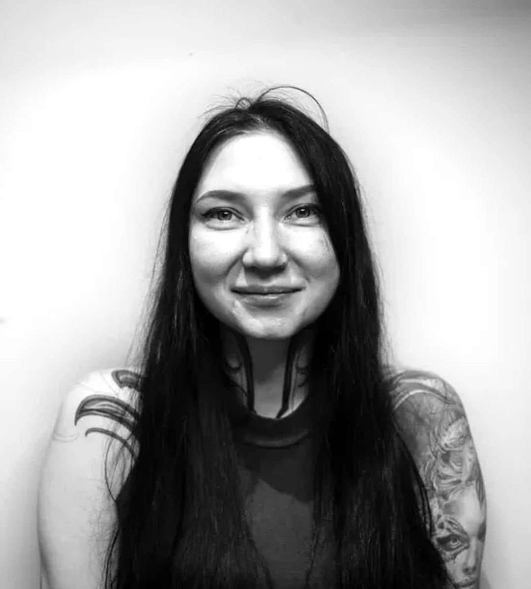 Woman with long dark hair, tattoos on neck and arms, smiling, looking at camera.