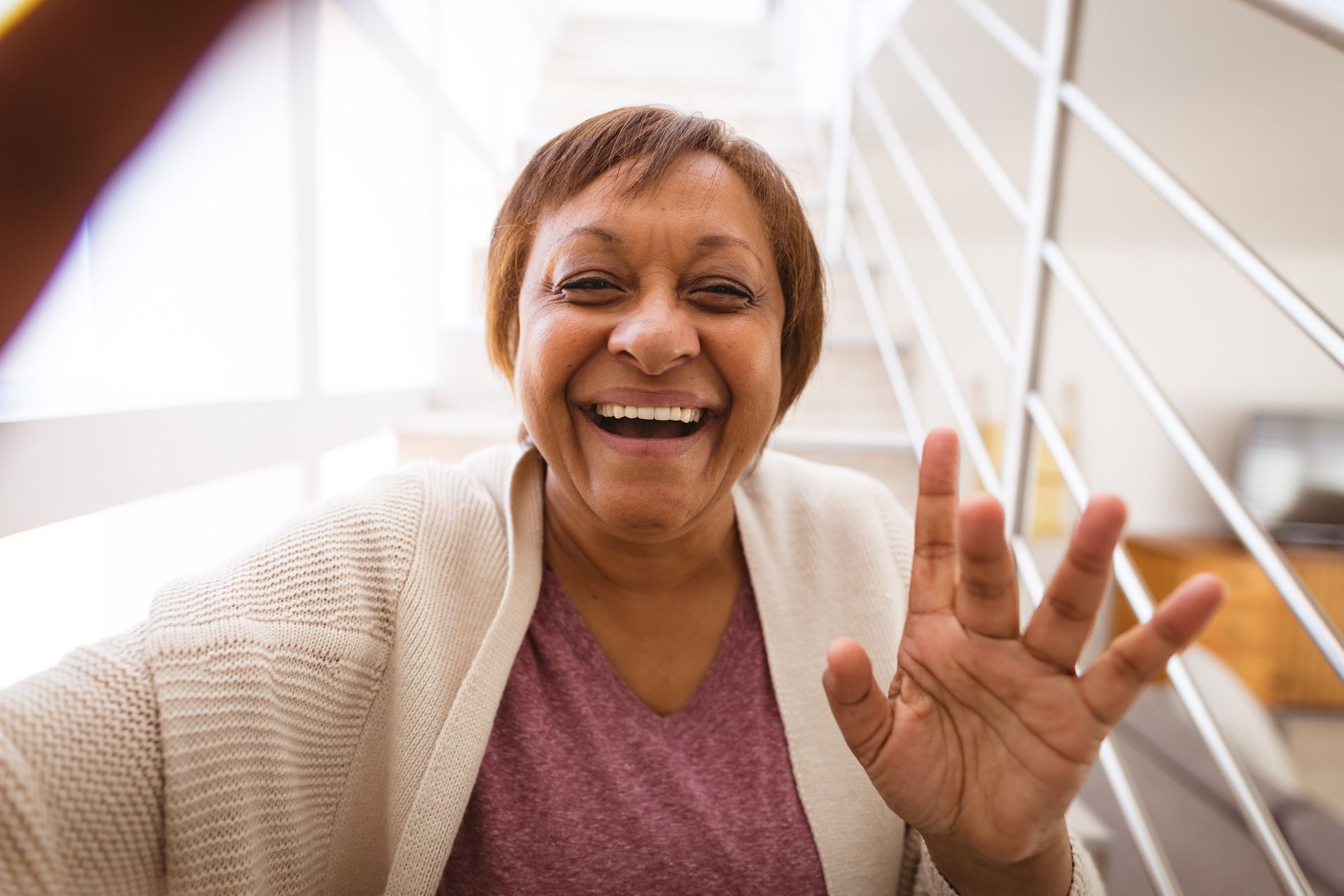 An elderly woman is waving at the camera while sitting on a set of stairs.