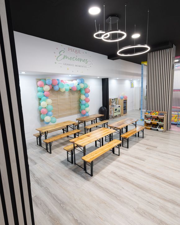 Aula luminosa con bancos de madera, pared de globos de colores y luces circulares en el techo.