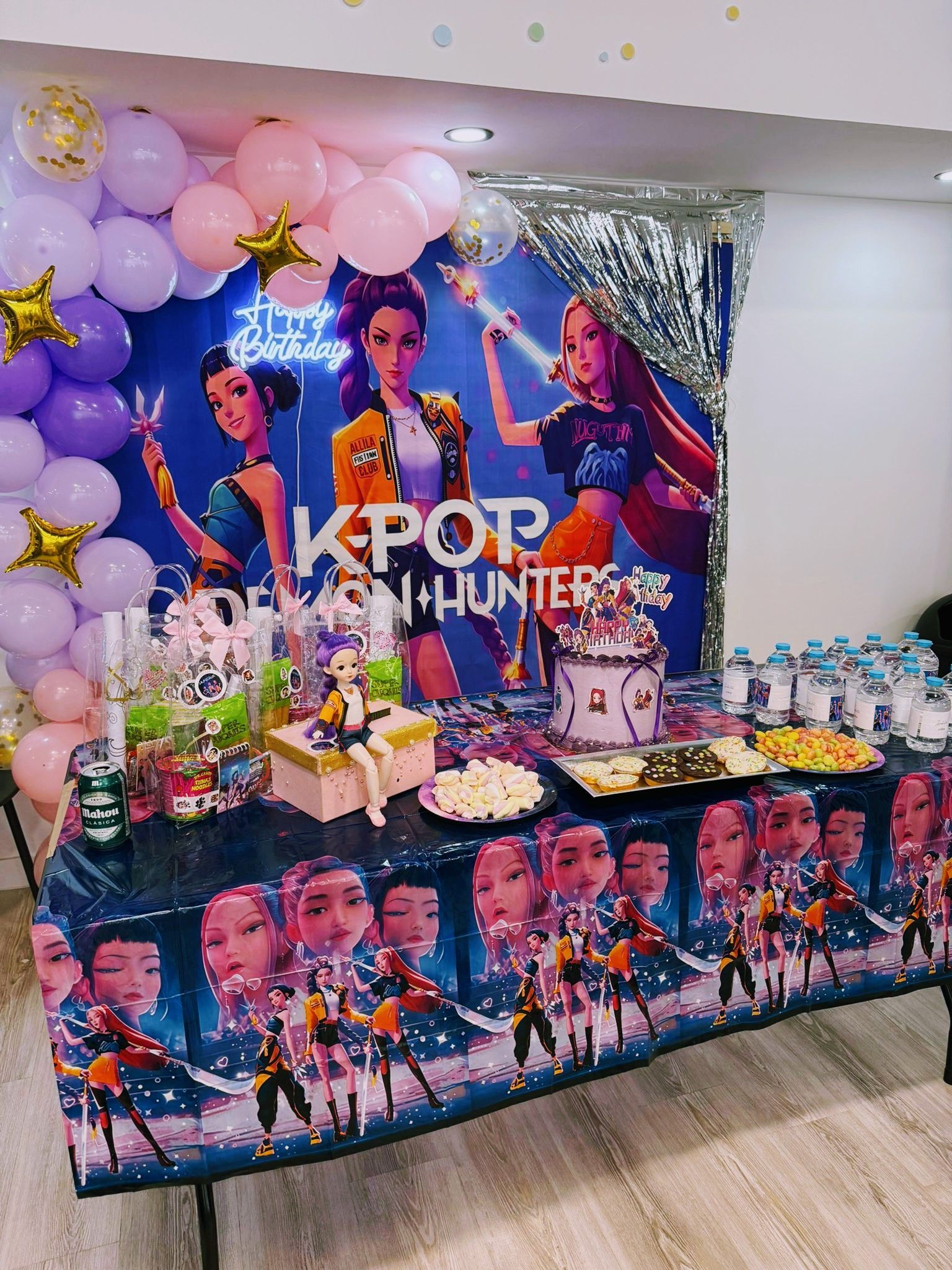 Mesa de postres para fiesta temática de K-pop con colorido fondo de personajes, globos, regalos y pasteles decorados.
