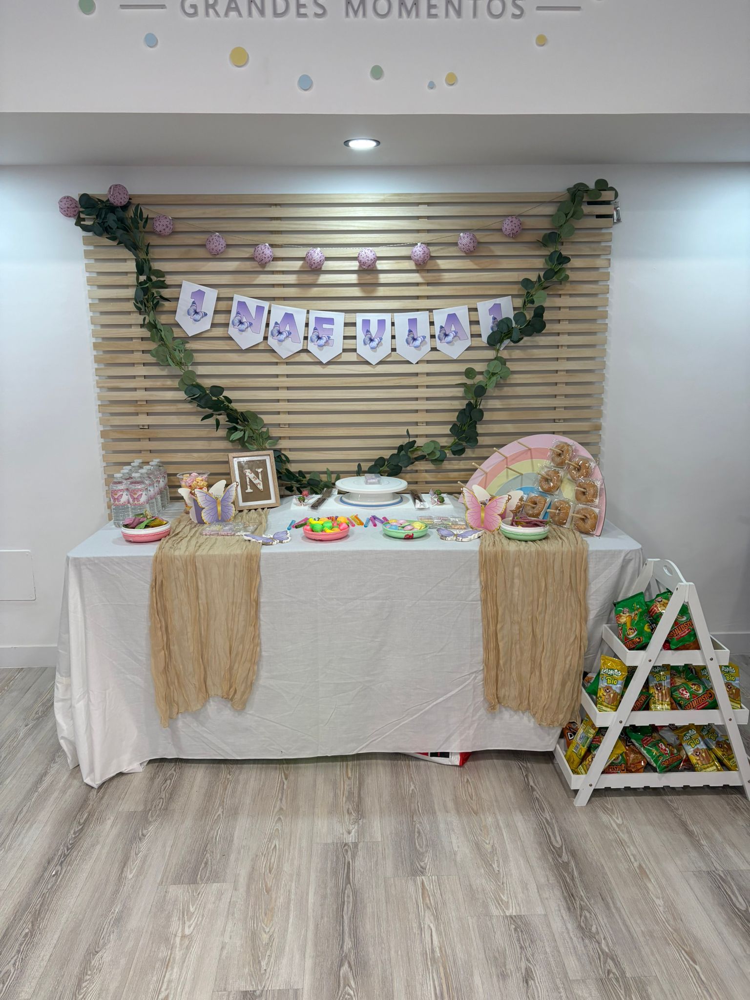 Mesa de postres decorada para fiestas con guirnaldas, banderines, globos y una pequeña escalera junto a un fondo blanco.