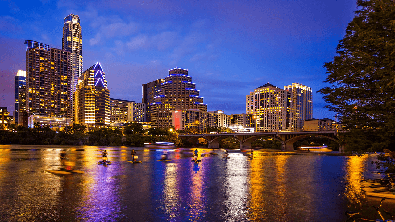 A group of people are paddling kayaks on a lake in front of a city skyline at night.